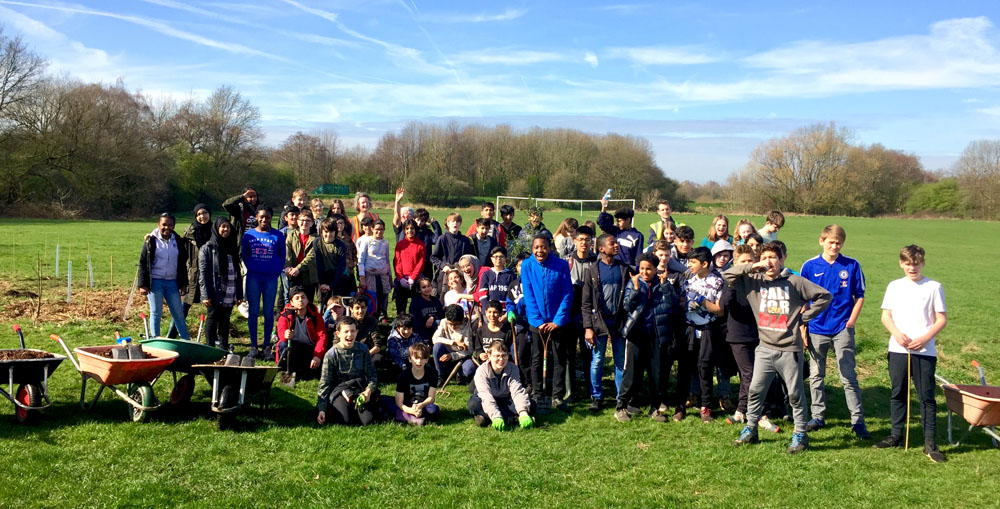 Large school group of young volunteers cheer for the camera at Turn Moss fields, some with wheelbarrows and spades for tree planting.